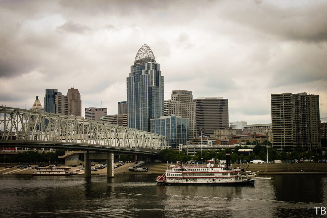 Riverboat and bridge with city skyline under cloudy sky.